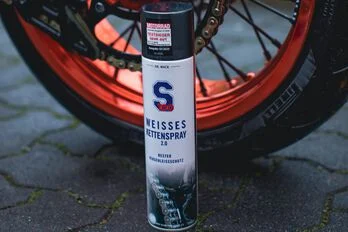 Sprühdose mit weißem Kettenspray vor einem Motorradreifen mit orangefarbener Felge auf einem gepflasterten Boden, bei der Motorradpflege mit S100 Produkt