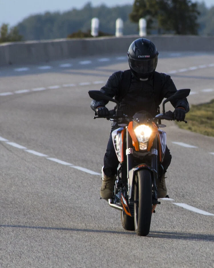 Motorradfahrer beim Motorradfahrsicherheitstraining mit schwarzer Schutzkleidung und Helm auf einer Landstraße, frontal fotografiert, mit eingeschaltetem Scheinwerfer.