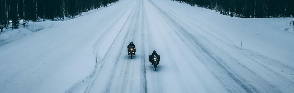 Zwei Motorradfahrer auf einer schneebedeckten Straße, umgeben von dichtem Wald bei winterlichen Bedingungen