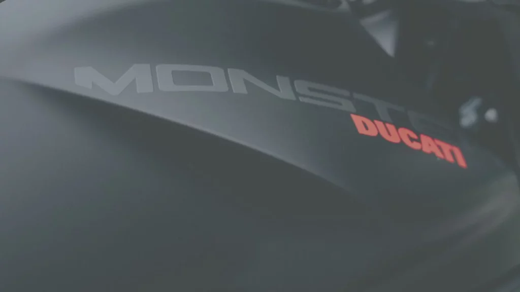 Nahaufnahme des Tanks einer Ducati Monster mit dem markanten Ducati-Logo in Rot und dem Monster-Schriftzug in Grau.