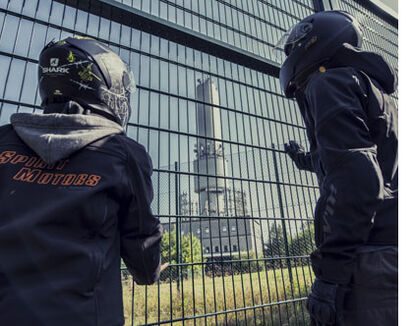 Zwei Personen mit Motorradhelmen blicken durch einen Zaun auf einen verlassenen Turm, ein markantes Ziel innerhalb eines Lost Places.